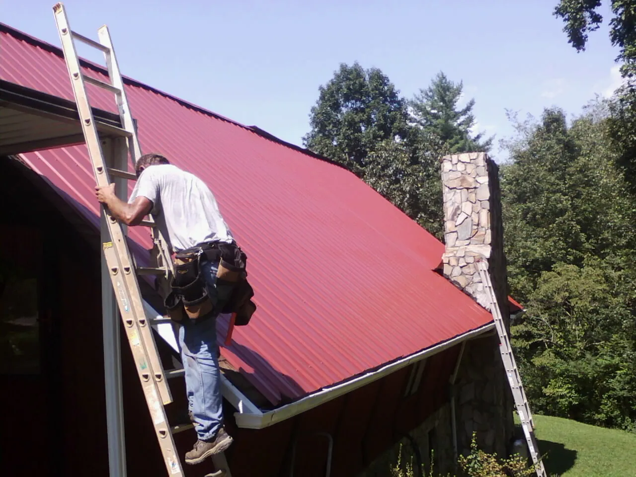 Roofer on a ladder beside a red metal roof and stone chimney — Soffit & Fascia Repair by Roofing Brothers in East Haddam